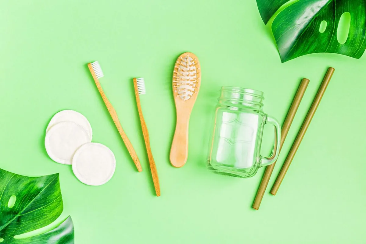 Bamboo toothbrushes, a wooden hairbrush, cotton pads and a glass jar on a green background